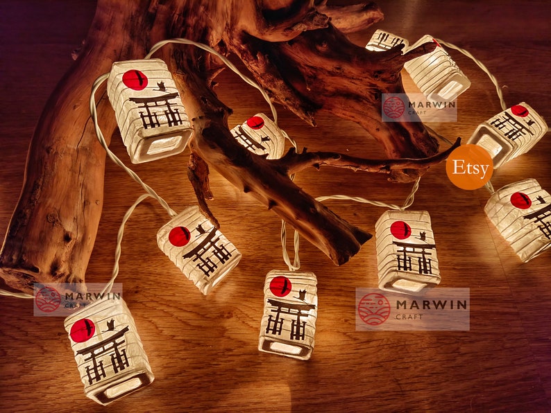 K&ouml;nnte beinhalten: Lichterkette aus wei&szlig;en Papierlaternen mit einer roten Sonne und schwarzen japanischen Schriftzeichen. Die Laternen sind beleuchtet und h&auml;ngen an einem Ast. Der Text "Marwin Craft" und "Etsy" ist im Bild sichtbar.