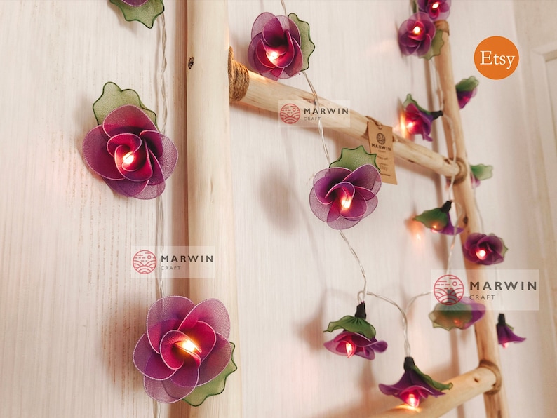 May include: Purple flower string lights with warm white LED bulbs. The lights are strung on a wire and are decorated with green leaves. The lights are hanging on a wooden ladder. The image also includes the text "Etsy" and "Marwin Craft".