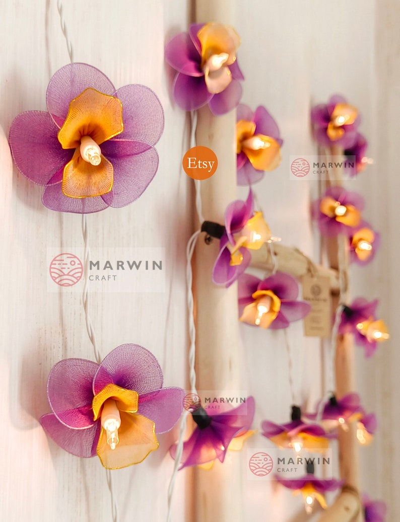 Puede incluir: Luces de cadena con bombillas en forma de flor de color morado y naranja. Las luces est&aacute;n enhebradas en un cable blanco y est&aacute;n encendidas. Las flores est&aacute;n hechas de tela y tienen un aspecto delicado y femenino. Las luces son perfectas para agregar un toque de fantas&iacute;a y romance a cualquier espacio.