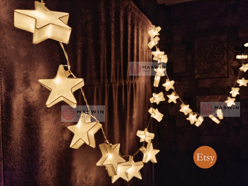 May include: A string of white paper star lights with warm white LED bulbs. The lights are strung on a clear wire and are hanging on a brown wall. The text "Marwin Craft" and "Etsy" are visible in the image.