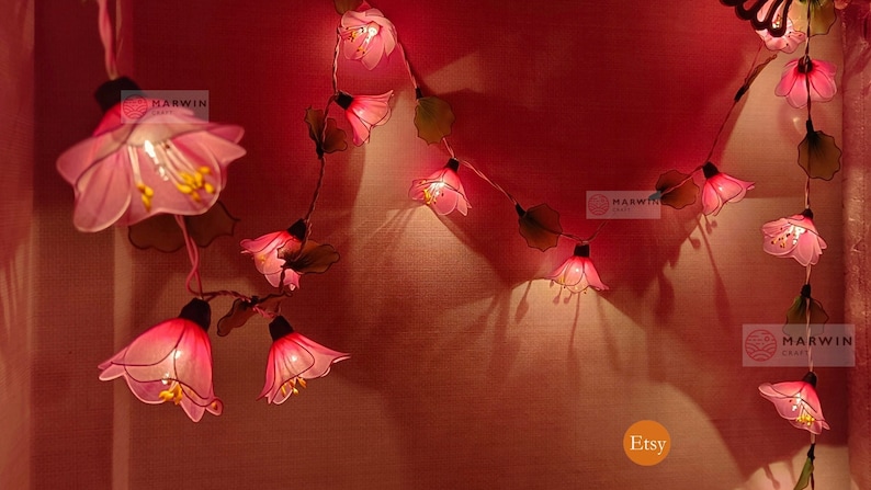 May include: A string of pink flower-shaped fairy lights with a warm glow. The lights are strung on a wire with green leaves. The lights are hanging on a red wall. The text "Etsy" and "Marwin Craft" are visible in the image.