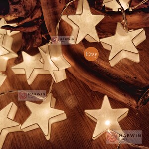 May include: String lights with white paper star-shaped lights. The lights are strung on a clear wire and are lit up. The lights are on a wooden surface. The text "Marwin Craft" and "Etsy" are visible in the image.