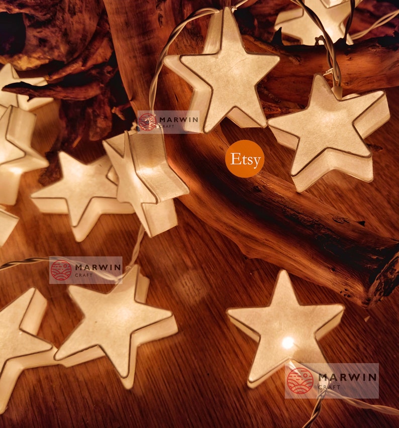May include: String lights with white paper star-shaped lights. The lights are strung on a clear wire and are lit up. The lights are on a wooden surface. The text "Marwin Craft" and "Etsy" are visible in the image.