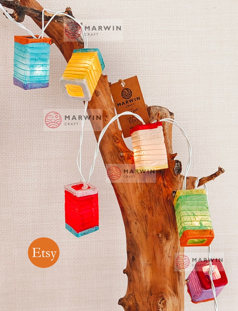 May include: A string of lights with paper lanterns in various colors, including blue, yellow, red, green, and purple. The lanterns are hanging from a branch of a tree. The lights are lit and the lanterns are glowing. The image also includes the text "Etsy" and "Marwin Craft".