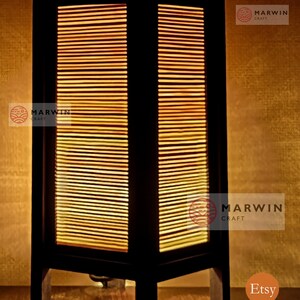 May include: A black wooden table lamp with a rectangular shade made of woven bamboo. The lamp is lit and casts a warm glow. The lamp is on a wooden surface. The text "Marwin Craft" is visible on the lamp.