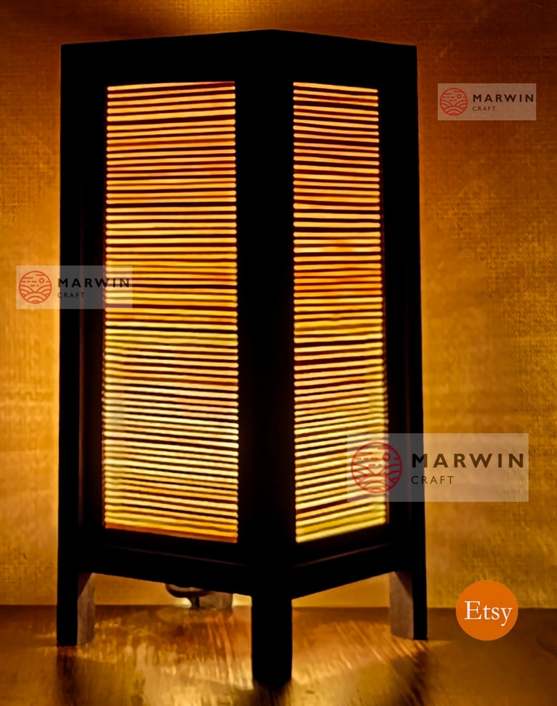 May include: A black wooden table lamp with a rectangular shade made of woven bamboo. The lamp is lit and casts a warm glow. The lamp is on a wooden surface. The text "Marwin Craft" is visible on the lamp.