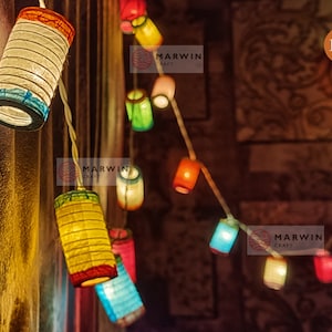May include: String of colorful paper lanterns, each with a unique shape and hue, including red, yellow, blue, and green. The lanterns are strung together on a white wire, creating a festive and decorative lighting display.