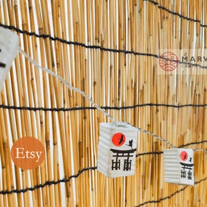 K&ouml;nnte beinhalten: Eine Girlande aus wei&szlig;en Papierlaternen mit einem roten Sonnen- und einem schwarzen Vogeldruck, die an einer Bambuswand h&auml;ngen. Die Laternen tragen die japanischen Schriftzeichen "Torii". Der Text "Etsy" und "Marwin Craft" ist auf dem Bild zu sehen.