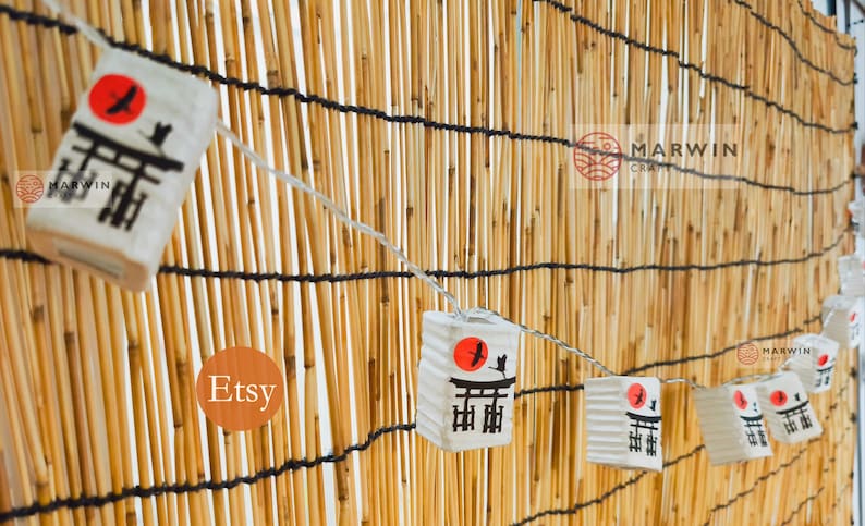 K&ouml;nnte beinhalten: Eine Girlande aus wei&szlig;en Papierlaternen mit einem roten Sonnen- und einem schwarzen Vogeldruck, die an einer Bambuswand h&auml;ngen. Die Laternen tragen die japanischen Schriftzeichen "Torii". Der Text "Etsy" und "Marwin Craft" ist auf dem Bild zu sehen.
