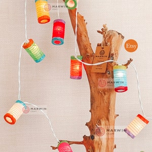 May include: A string of colorful paper lanterns, each with a unique design and hue, are strung along a white wire. The lanterns are illuminated, casting a warm glow. The string of lights is draped over a piece of driftwood, creating a decorative display.