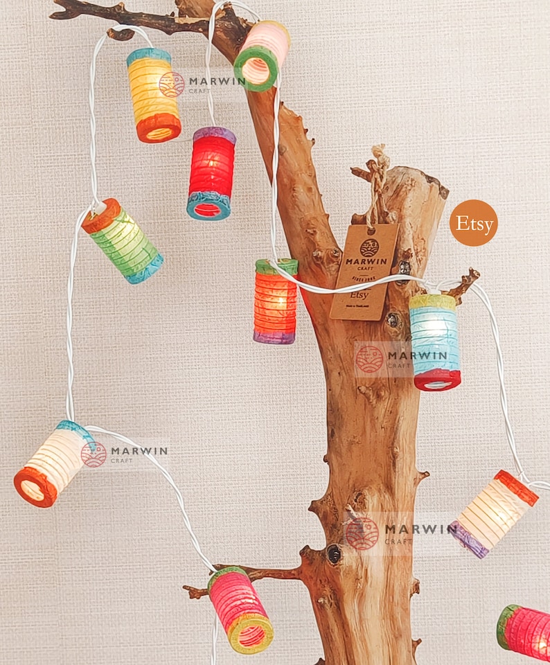 May include: A string of colorful paper lanterns, each with a unique design and hue, are strung along a white wire. The lanterns are illuminated, casting a warm glow. The string of lights is draped over a piece of driftwood, creating a decorative display.