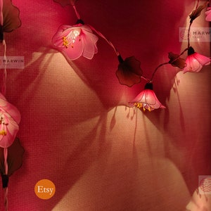 May include: A string of pink flower lights with brown leaves, lit up and hanging on a pink wall. The lights are labelled "Marwin Craft" and "Etsy".