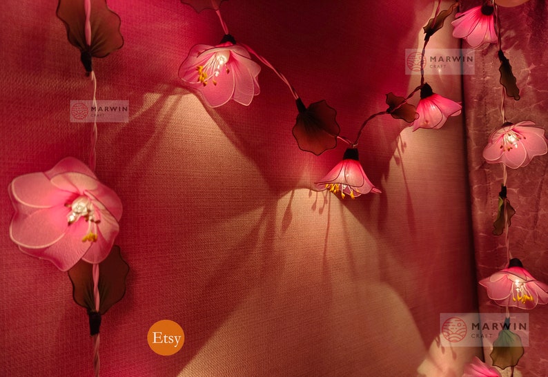 May include: A string of pink flower lights with brown leaves, lit up and hanging on a pink wall. The lights are labelled "Marwin Craft" and "Etsy".