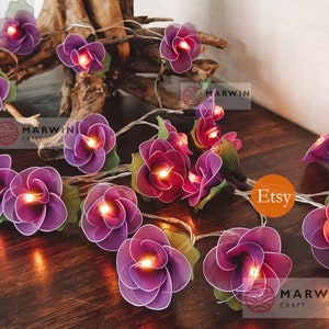 May include: Purple flower shaped fairy lights with warm white LED bulbs. The lights are strung on a clear wire and are perfect for adding a touch of whimsy to any space. The lights are handmade and feature a delicate, layered design. The lights are perfect for adding a touch of whimsy to any space.  Etsy Marwin Craft.