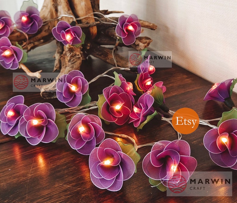 May include: Purple flower shaped fairy lights with warm white LED bulbs. The lights are strung on a clear wire and are perfect for adding a touch of whimsy to any space. The lights are handmade and feature a delicate, layered design. The lights are perfect for adding a touch of whimsy to any space.  Etsy Marwin Craft.