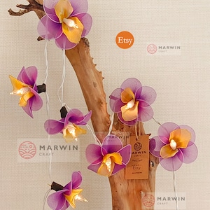 Puede incluir: Una cadena de luces con bombillas en forma de flores moradas y amarillas. Las luces est&aacute;n enhebradas en una rama y las flores est&aacute;n hechas de tela. Las luces est&aacute;n encendidas y las bombillas brillan. La imagen tambi&eacute;n incluye una peque&ntilde;a etiqueta marr&oacute;n con el texto "Marwin Craft Etsy" en ella.