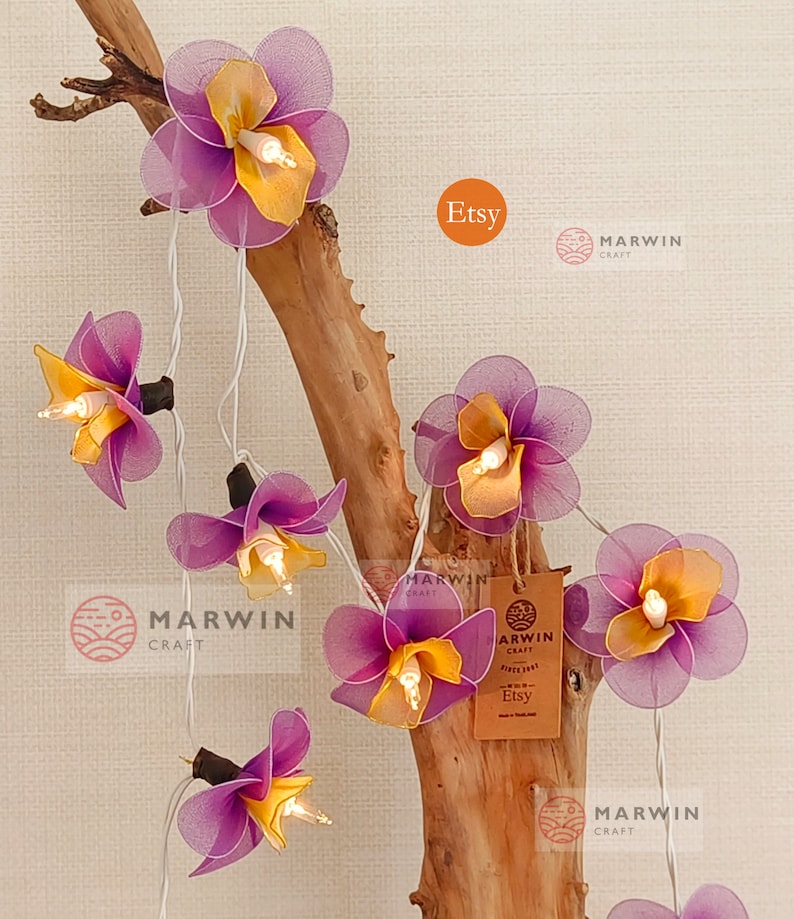 Puede incluir: Una cadena de luces con bombillas en forma de flores moradas y amarillas. Las luces est&aacute;n enhebradas en una rama y las flores est&aacute;n hechas de tela. Las luces est&aacute;n encendidas y las bombillas brillan. La imagen tambi&eacute;n incluye una peque&ntilde;a etiqueta marr&oacute;n con el texto "Marwin Craft Etsy" en ella.