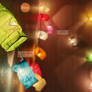 May include: A string of colorful paper lanterns with warm white lights. The lanterns are made of thin, crinkled paper and are strung together with a clear wire. The lanterns are hanging from a wall and are casting a warm glow on the surrounding area. The text "Etsy" and "Marwin Craft" are visible in the image.