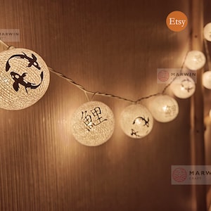 May include: A string of white cotton ball lights with different designs, including fish and Chinese characters. The lights are strung on a wire and are lit up, casting a warm glow.