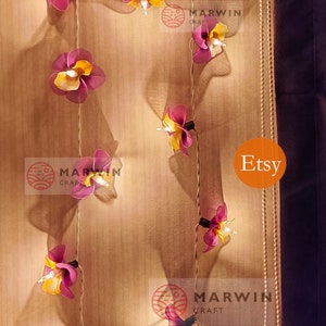 Puede incluir: Una cadena de luces con bombillas en forma de flor de color morado y amarillo. Las luces est&aacute;n enhebradas en un cable blanco y est&aacute;n colgadas en una pared beige. El texto "Marwin Craft" y "Etsy" es visible en la imagen.