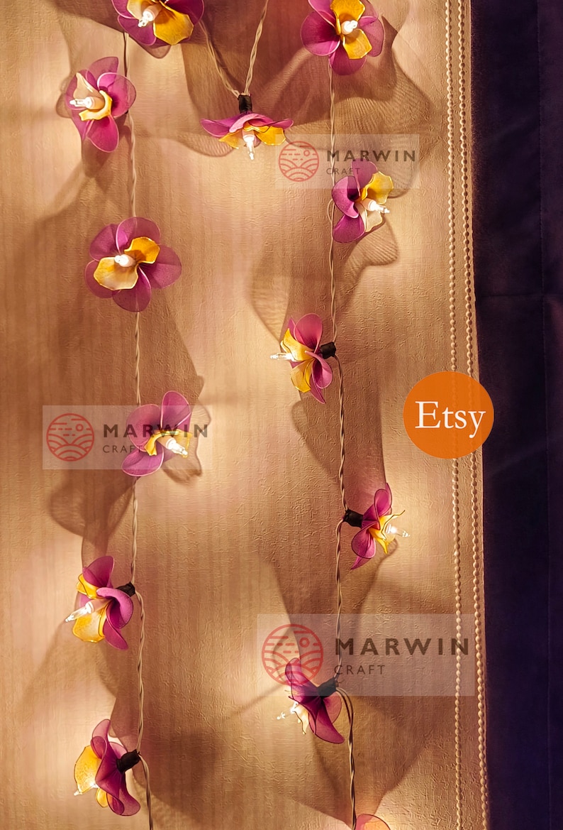 Puede incluir: Una cadena de luces con bombillas en forma de flor de color morado y amarillo. Las luces est&aacute;n enhebradas en un cable blanco y est&aacute;n colgadas en una pared beige. El texto "Marwin Craft" y "Etsy" es visible en la imagen.