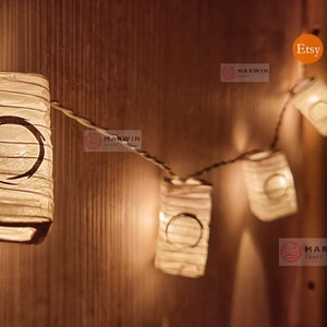 May include: A string of white paper lanterns with a black circle design, lit with warm white LED lights. The lanterns are hanging on a wooden wall with the word "Etsy" and the logo "Marwin Craft" visible.