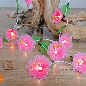 May include: A string of fairy lights with pink fabric flowers. The lights are strung on a white wire and have small white bulbs. The flowers are made of a delicate fabric and have a pink and white colour scheme. The lights are perfect for adding a touch of whimsy to any room.