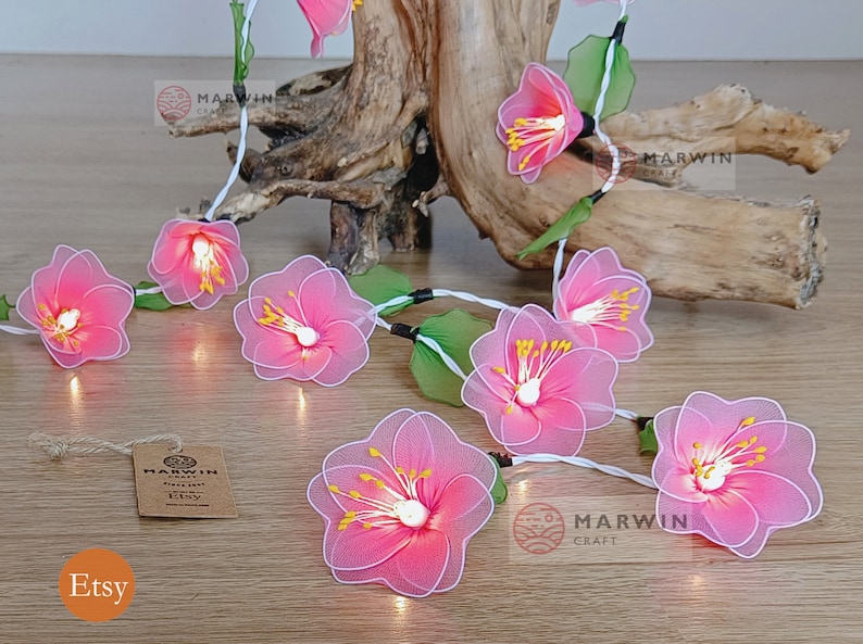 May include: A string of fairy lights with pink fabric flowers. The lights are strung on a white wire and have small white bulbs. The flowers are made of a delicate fabric and have a pink and white colour scheme. The lights are perfect for adding a touch of whimsy to any room.
