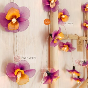 Puede incluir: Una cadena de luces con bombillas en forma de flor de color morado y naranja. Las luces est&aacute;n enhebradas en un cable blanco y est&aacute;n colgadas de una escalera de madera. El texto "Etsy" y "Marwin Craft" es visible en la imagen.