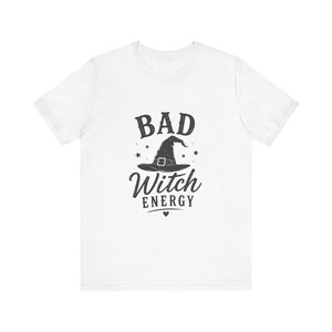 May include: A white short-sleeved t-shirt featuring a black graphic print on the chest. The design includes a witch hat in the center, surrounded by small stars, with the text "BAD" above the hat and "Witch ENERGY" below it, finished with a small heart.