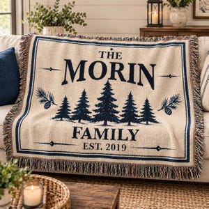 Puede incluir: Una manta tejida en color crema con borde y flecos azul oscuro. El centro presenta pinos azul oscuro, piñas y el texto: THE MORIN FAMILY EST. 2019. El diseño está enmarcado por una fina línea azul oscuro con detalles decorativos.