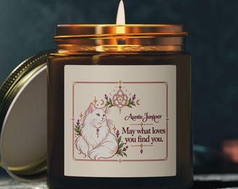 May include: A brown glass candle jar with a gold lid and a white label featuring a cat illustration, floral accents, and the text Auntie Juniper May what loves you find you. The candle has a lit wick and a warm glow from within.