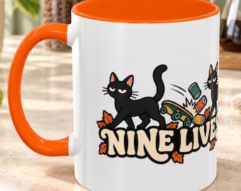 May include: A white ceramic mug with a bright orange handle and interior. The mug features a graphic of two black cats, a skateboard, and scattered cans, with the text NINE LIVES in a retro font surrounded by orange autumn leaves. Capacity is 325 ml.
