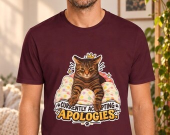May include: A maroon short-sleeved t-shirt featuring a graphic of a brown tabby cat wearing a small gold crown, resting on a polka-dot pillow. Below the cat, the text reads "CURRENTLY ACCEPTING APOLOGIES" in bold yellow and white lettering.