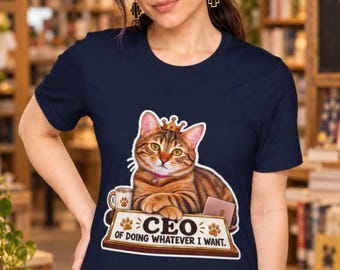 May include: A navy blue short-sleeve t-shirt featuring a graphic of a brown tabby cat wearing a gold crown. The cat rests its paws on a desk nameplate that reads "CEO OF DOING WHATEVER I WANT" with paw print accents, a coffee mug, and a laptop.