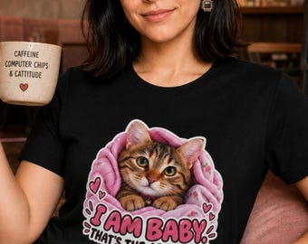 May include: A black t-shirt featuring a graphic of a brown tabby cat nestled in a pink blanket with the text "I AM BABY THAT'S THE ARGUMENT" and small hearts. A beige mug with the text "CAFFEINE COMPUTER CHIPS & CATTITUDE" is also visible.