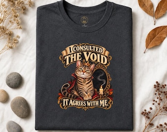 May include: A dark grey t-shirt featuring a graphic of a tabby cat wearing a crown and red cape, sitting before a mirror with a lit candle. The text reads I CONSULTED THE VOID IT AGREES WITH ME, framed by an ornate gold-coloured border design.