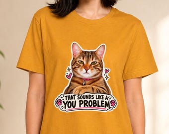 May include: A mustard yellow t-shirt featuring a graphic of a brown tabby cat wearing a pink collar with a bell. The cat is centred above the text "THAT SOUNDS LIKE A YOU PROBLEM" in black letters, accented by pink hearts, paw prints, and sparkles.