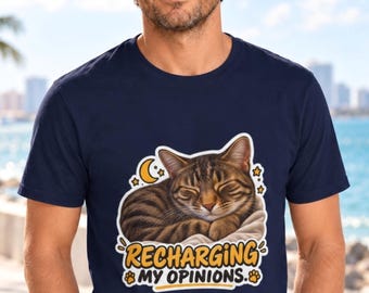 May include: A navy blue short-sleeved t-shirt featuring a graphic of a sleeping brown tabby cat on a white blanket. Above the cat is a yellow crescent moon and stars. Below the cat, the text reads RECHARGING MY OPINIONS in yellow letters.