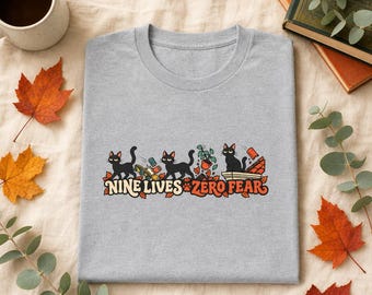 May include: A heather grey t-shirt featuring a graphic of three black cats with the text "NINE LIVES ZERO FEAR" in orange and brown. The illustration includes cats knocking over items, a potted plant, and scattered autumn leaves.