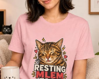 May include: A light pink t-shirt featuring a graphic of a brown tabby cat with its eyes closed and tongue sticking out. The text below the cat reads "RESTING MLEM FACE" in bold letters, surrounded by small red hearts and yellow star accents.