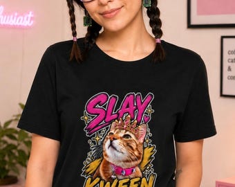 May include: A black t-shirt featuring a graphic of a ginger cat wearing a pink crown and a pink collar. The text SLAY is printed in large pink letters above the cat, and the word KWEEN is printed in large yellow letters below the cat.