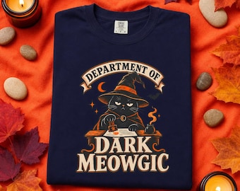 May include: A navy blue crew neck t-shirt featuring a graphic of a black cat wearing a witch hat and cape, sitting at a desk with a stamp. The text reads Department of Dark Meowgic. The shirt is made of cotton and has a size large label.