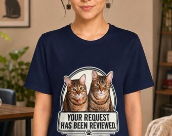 May include: A navy blue short-sleeved t-shirt featuring a graphic of two brown tabby cats. Below the cats, the text reads: YOUR REQUEST HAS BEEN REVIEWED. The answer is still no. A small paw print icon is positioned between the two lines of text.