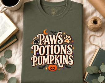 May include: An olive green t-shirt featuring a graphic print with the text Paws Potions Pumpkins in a cream font. The design includes a black cat, a paw print, a crescent moon, bats, and an orange jack-o'-lantern, accented with small colourful stars.