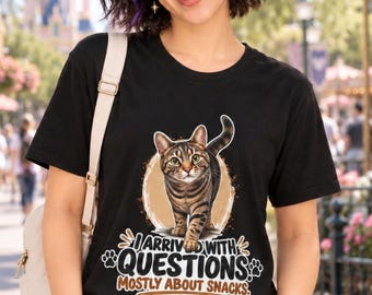 May include: A black short-sleeved t-shirt featuring a graphic of a brown tabby cat walking forward. Below the cat, the text reads: I ARRIVED WITH QUESTIONS MOSTLY ABOUT SNACKS, accompanied by small paw print icons and a circular background design.