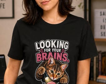 May include: A black short-sleeved t-shirt featuring a graphic of a brown tabby cat holding a flashlight and looking at a small pink brain. The text above the cat reads LOOKING FOR YOUR BRAINS in white and pink block lettering with a distressed effect.