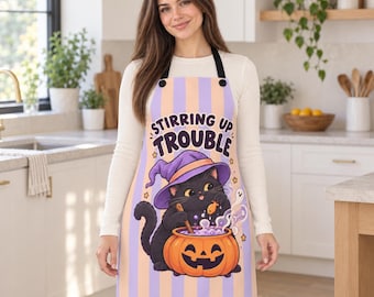 May include: A kitchen apron featuring vertical light purple and orange stripes. The centre graphic shows a black cat wearing a purple witch hat, stirring a bubbling orange pumpkin cauldron. The text reads STIRRING UP TROUBLE.