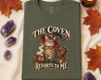 May include: An olive green t-shirt featuring a graphic of a tabby cat wearing a witch hat, sitting on a book next to a steaming cauldron. The text reads THE COVEN REPORTS TO ME in a decorative font. The design includes autumn leaves and a pentagram.