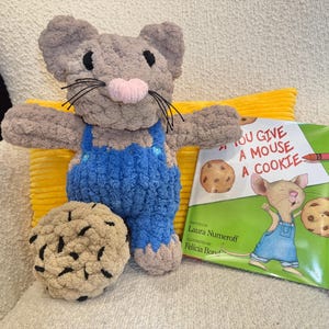 May include: A crocheted plush mouse with tan fur, a pink nose, and black whiskers, wearing blue overalls with small buttons. Beside it is a round, tan crocheted cookie with black speckles and a copy of the book If You Give a Mouse a Cookie.